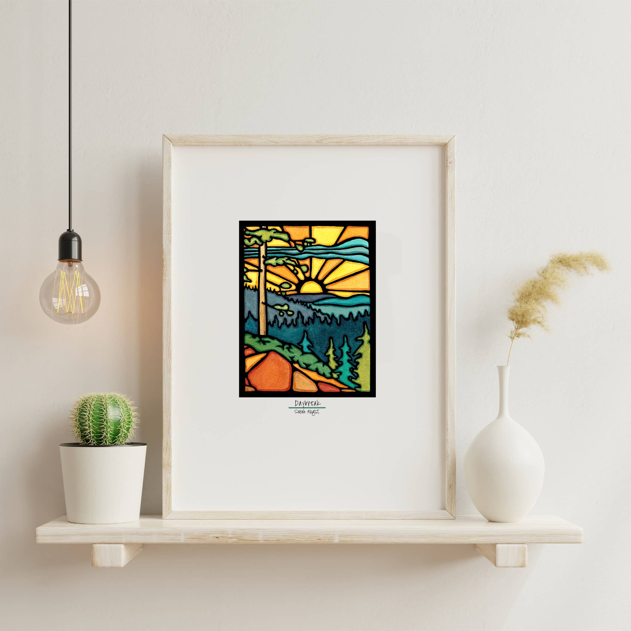 The "Daybreak: Print 11"x14" by Sarah Angst" framed artwork, featuring a stained-glass-style landscape of trees, hills, and a rising sun, sits on a white shelf near a cactus, a vase, and a hanging bulb.