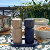 The Shakers: Salt and Pepper Grinder Set from Sweden, with sleek Scandinavian design, sits on a tray atop an outdoor table surrounded by food, lush plants, and a scenic backdrop.