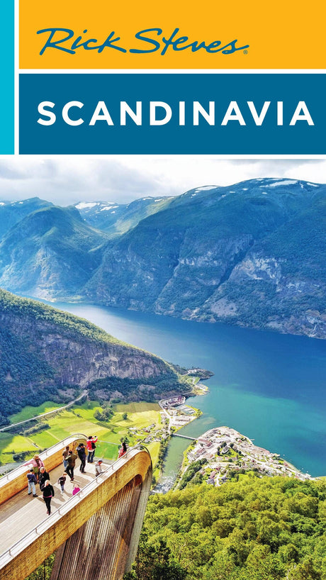 A group stands on a wooden platform overlooking a fjord, mountains, and village below—a view straight from the "Rick Steves Scandinavia" book—beneath a partly cloudy sky.