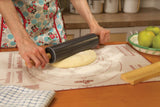 A person rolls out dough on a pastry mat using the Black Marble Rolling Pin with Wood Base; a checkered towel and green apples rest on the wooden counter nearby.