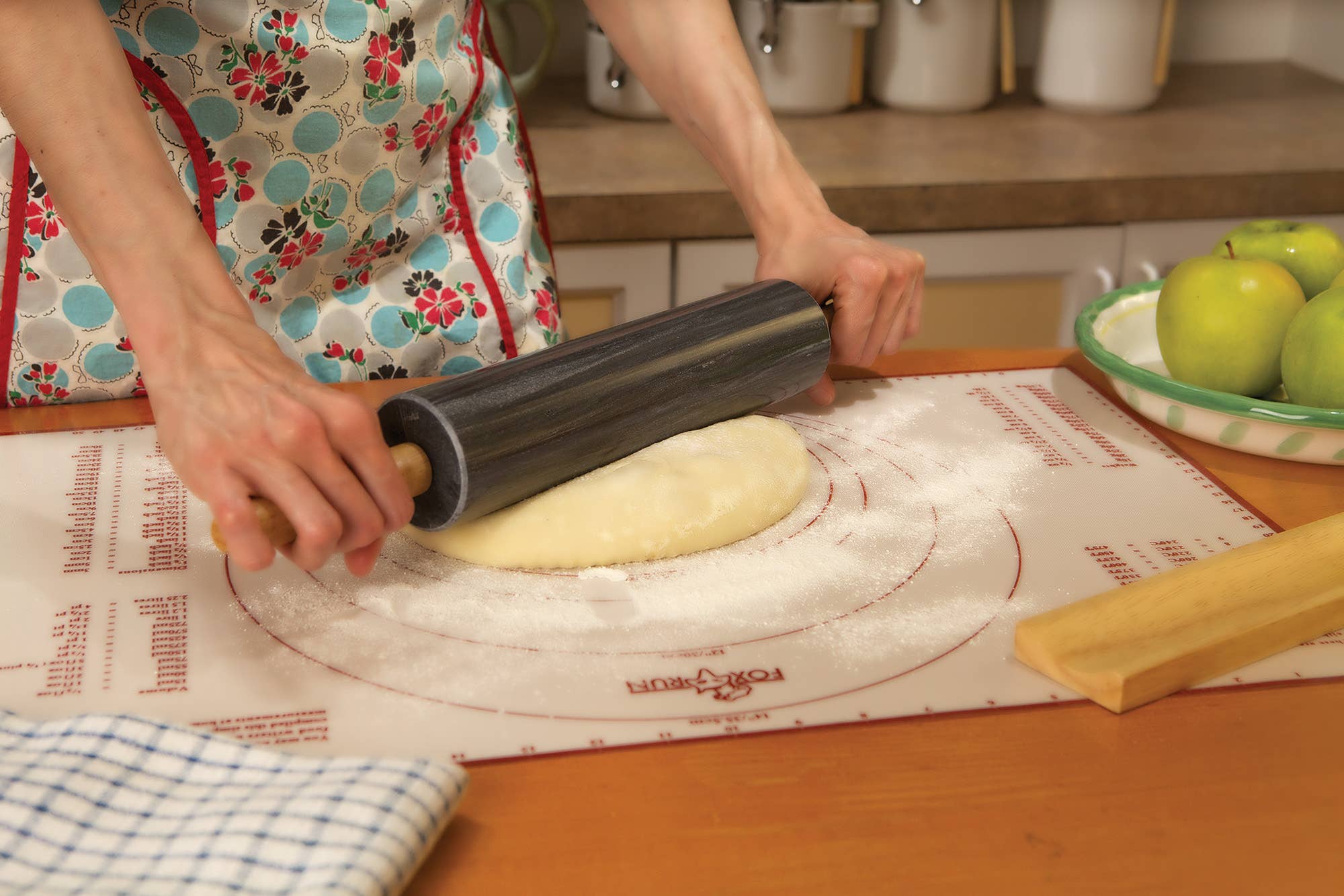 A person is rolling out dough on a floured surface with the sleek Marble Rolling Pin in Black, which features wooden handles, next to green apples and a folded kitchen towel.