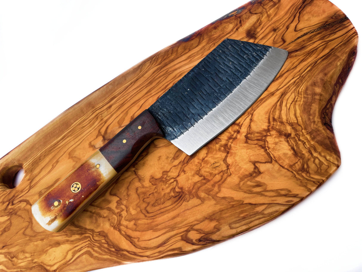 The Handmade Carbon Steel Cleaver for Kitchen & Outdoor Use, featuring a textured black blade and wooden handle, rests on a polished wooden cutting board.
