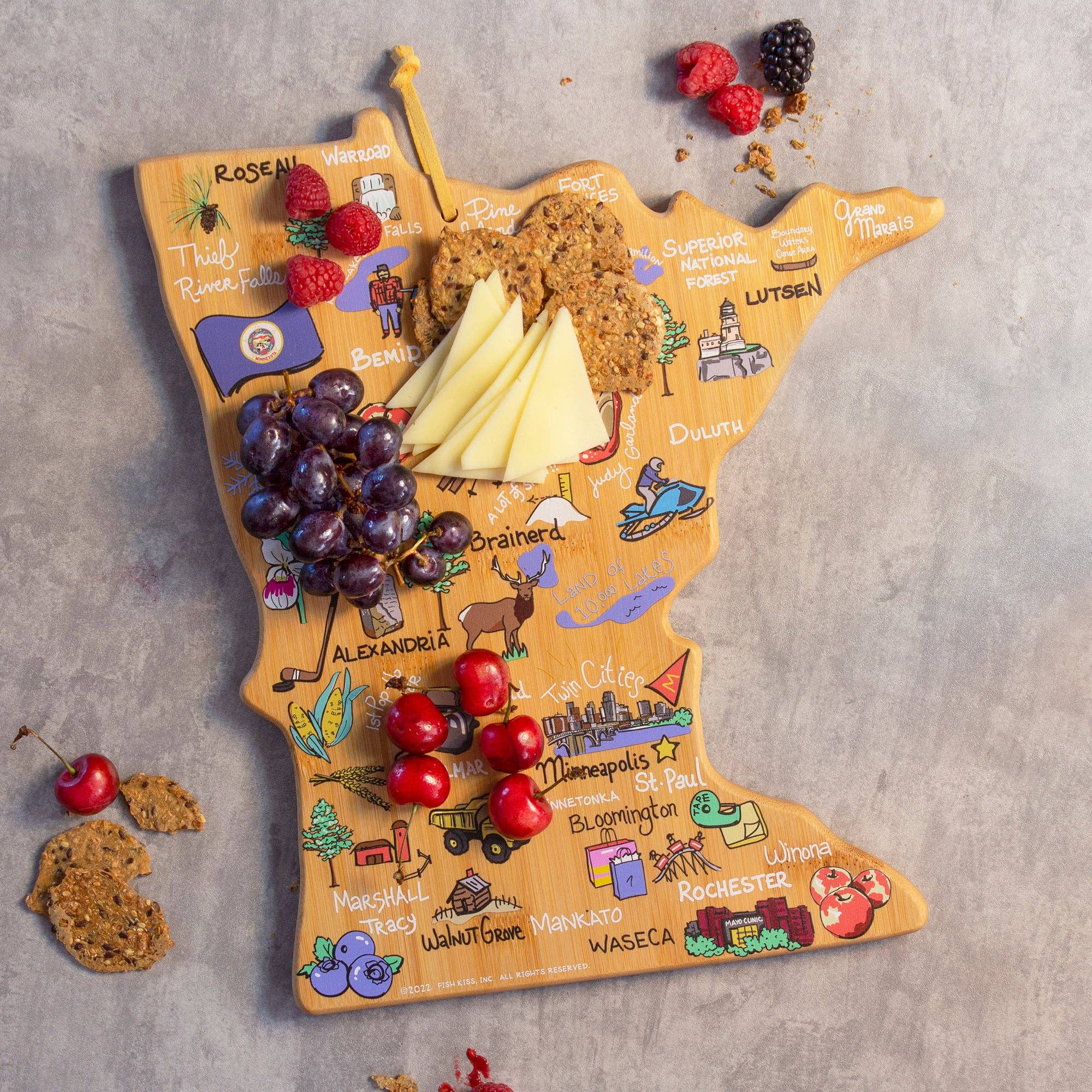 A durable bamboo cutting board, designed to resemble the state of Minnesota and featuring detailed illustrations and town names, serves as an elegant backdrop for grapes, cheese slices, cherries, and crackers. This functional and decorative piece is known as the "Minnesota Cutting Board with Artwork by Fish Kiss™.