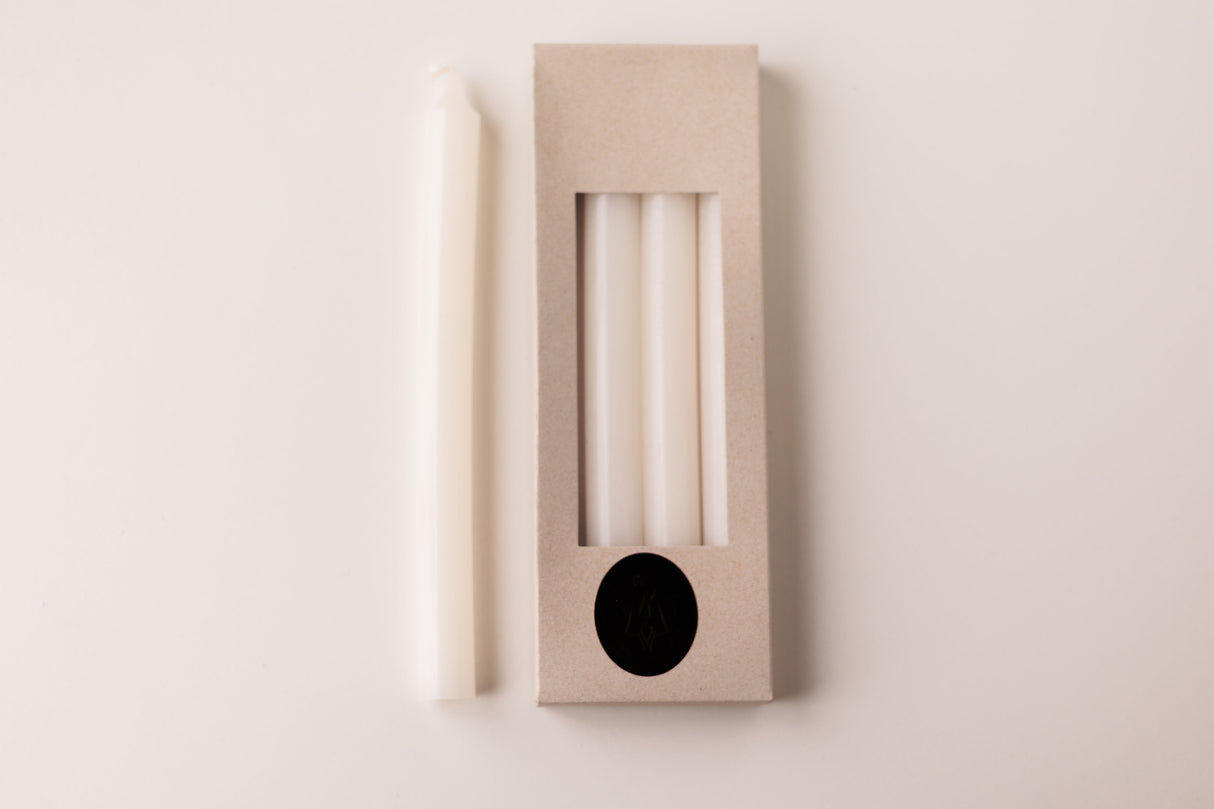 A white taper candle rests beside a box labeled "White Crown 10\" Candles 4 Pack from Logum Kloster, Denmark," showing three more Danish candles through a clear window in the packaging.