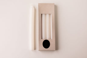 A white taper candle rests beside a box labeled "White Crown 10\" Candles 4 Pack from Logum Kloster, Denmark," showing three more Danish candles through a clear window in the packaging.