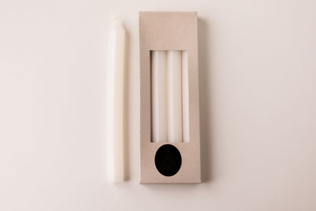 A white taper candle rests beside a box labeled "White Crown 10\" Candles 4 Pack from Logum Kloster, Denmark," showing three more Danish candles through a clear window in the packaging.
