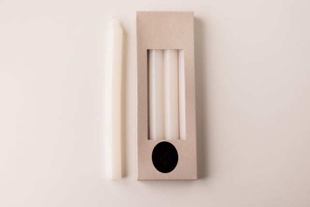A white taper candle rests beside a box labeled "White Crown 10\" Candles 4 Pack from Logum Kloster, Denmark," showing three more Danish candles through a clear window in the packaging.