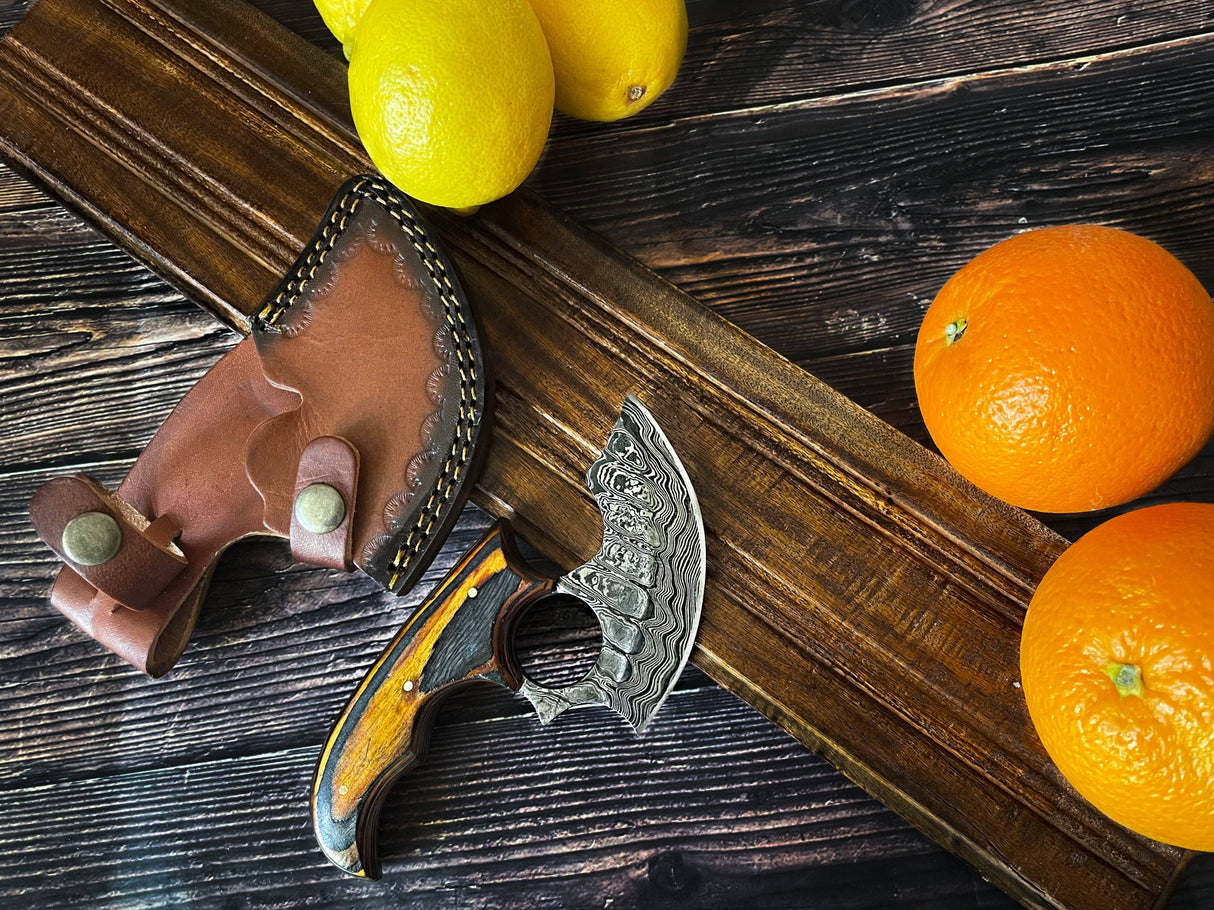 The Knife: Alaskan Ulu Style Blade with a high carbon Damascus steel blade and wooden handle rests by its leather sheath, surrounded by lemons and oranges on a dark wooden surface.