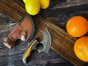 The Knife: Alaskan Ulu Style Blade with a high carbon Damascus steel blade and wooden handle rests by its leather sheath, surrounded by lemons and oranges on a dark wooden surface.