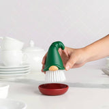 A hand holds a gnome dish brush with a green hat above a red dish, with stacked white dishes visible in the background.