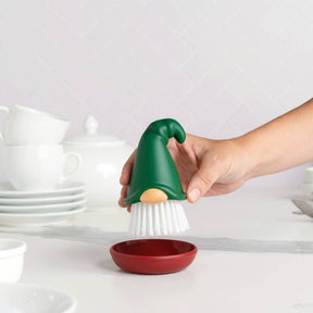 A hand holds a gnome dish brush with a green hat above a red dish, with stacked white dishes visible in the background.