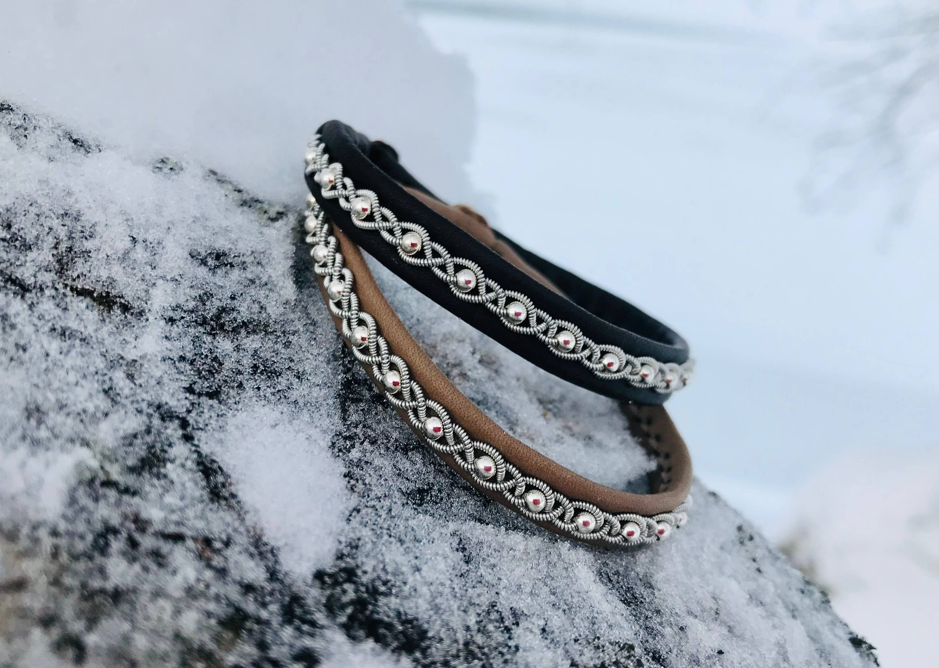 A handcrafted Sami Lapland Bracelet (#27) in black, made with reindeer hide and pewter silver beads, rests on a snow-covered rock outdoors.