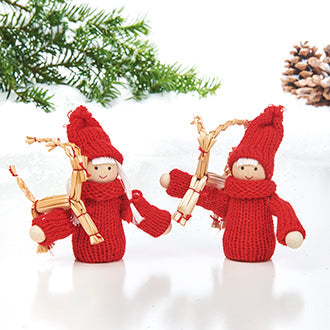 Two small wooden dolls, inspired by Scandinavian Pixies, wear red knit outfits and hold twigs. A pine branch and pinecone add to the festive display on a white background, embodying the charm of Swedish Christmas Decorations. .