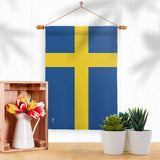 A Sweden Garden Flag made of weather-resistant fabric hangs above a wooden crate with a vase of flowers and potted succulents, bringing Scandinavian charm to the white space.
