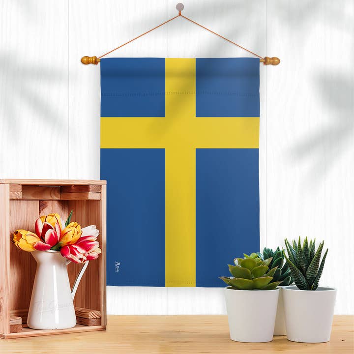 A Sweden Garden Flag made of weather-resistant fabric hangs above a wooden crate with a vase of flowers and potted succulents, bringing Scandinavian charm to the white space.