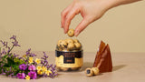 A hand picks up a Crispy Caramel Danish Chocolate Coated Licorice candy (Lakrids by Bulow, 125g) from a jar, with flowers and a caramel shard nearby.