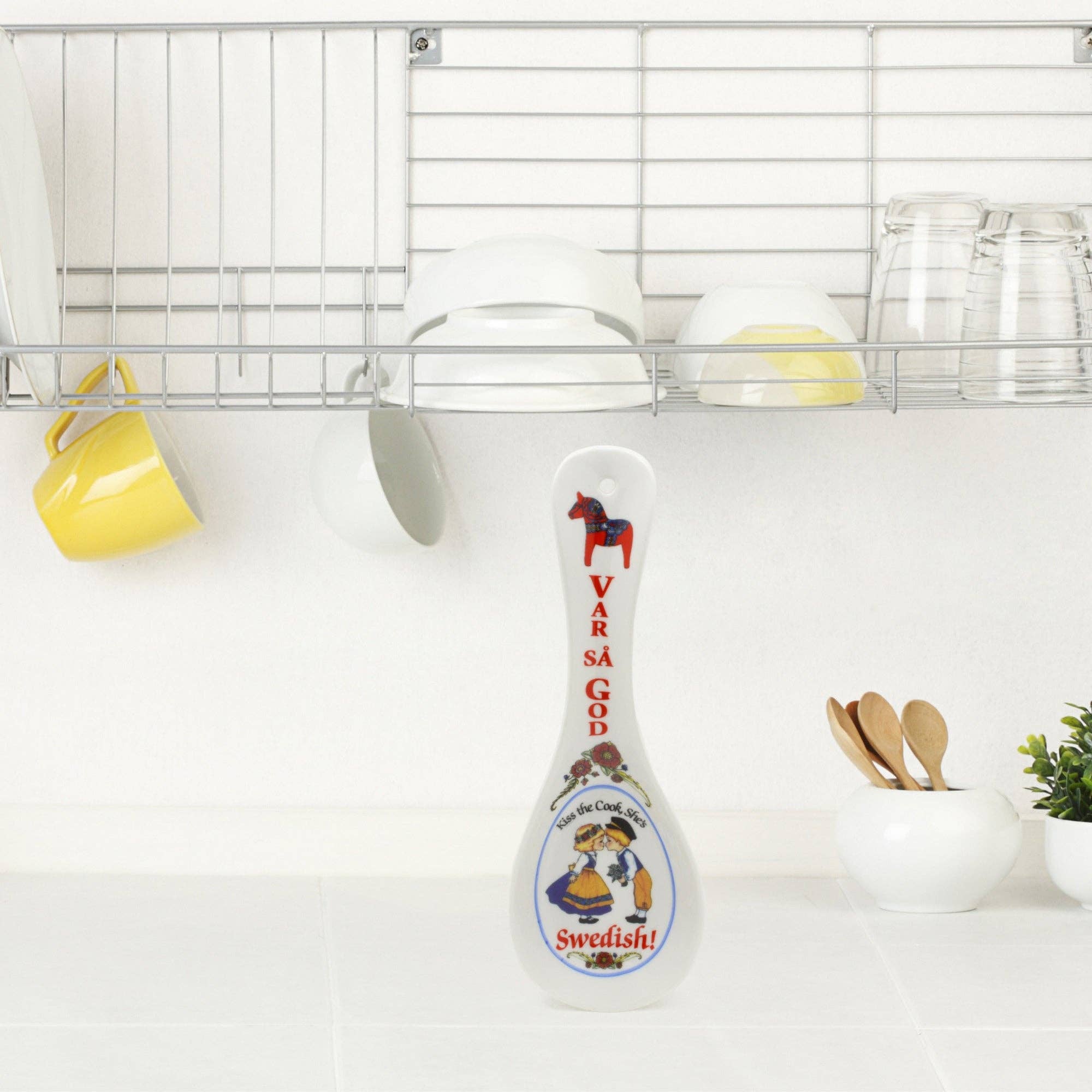 The Spoon Rest: Swedish Gift Var Sa God, adorned with red text and illustrations, stands elegantly on a kitchen countertop beside dishes and cups on the shelf above, serving as both a functional tool and charming kitchen decor.