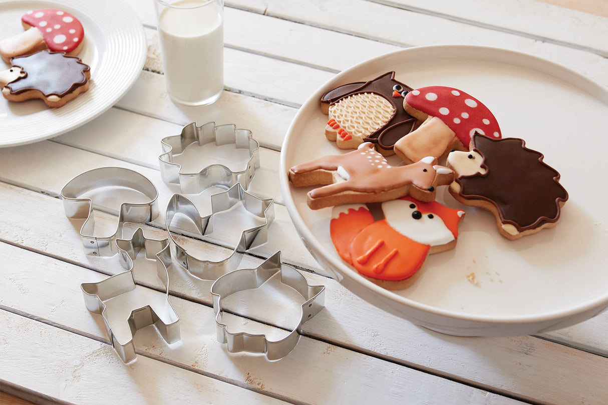 Artfully arranged on a plate are decorative cookies in the shapes of animals and mushrooms, which are encircled by the Cookie Cutters: Woodland Animal Set of 5, crafted from stainless steel, with a glass of milk placed on the white wooden table.