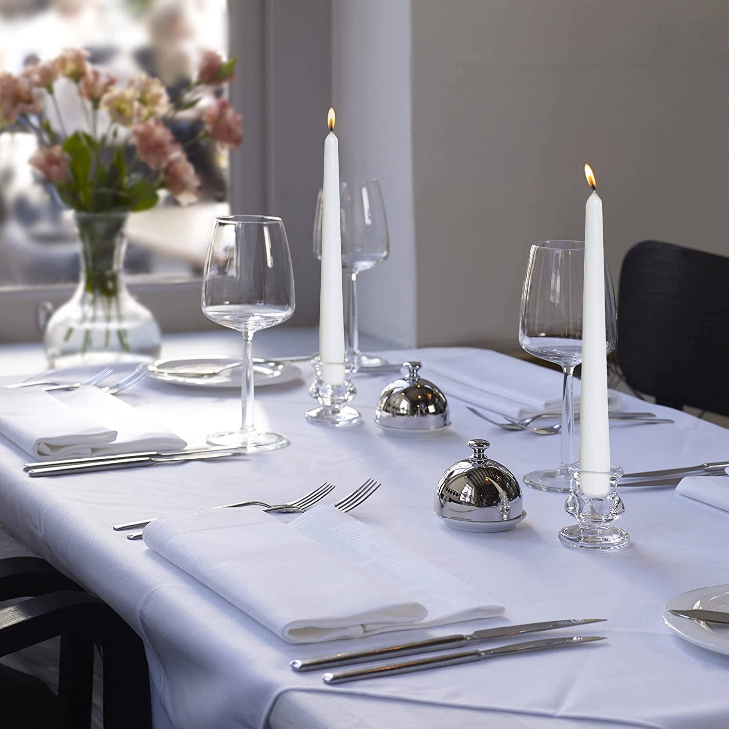 A formal dining table setting features high-quality, unscented dripless candles from the White Tall 10" Taper 4 Pack casting a warm glow. Empty wine glasses, silver cutlery, and white napkins rest elegantly on a white tablecloth. A vase of flowers graces the background.