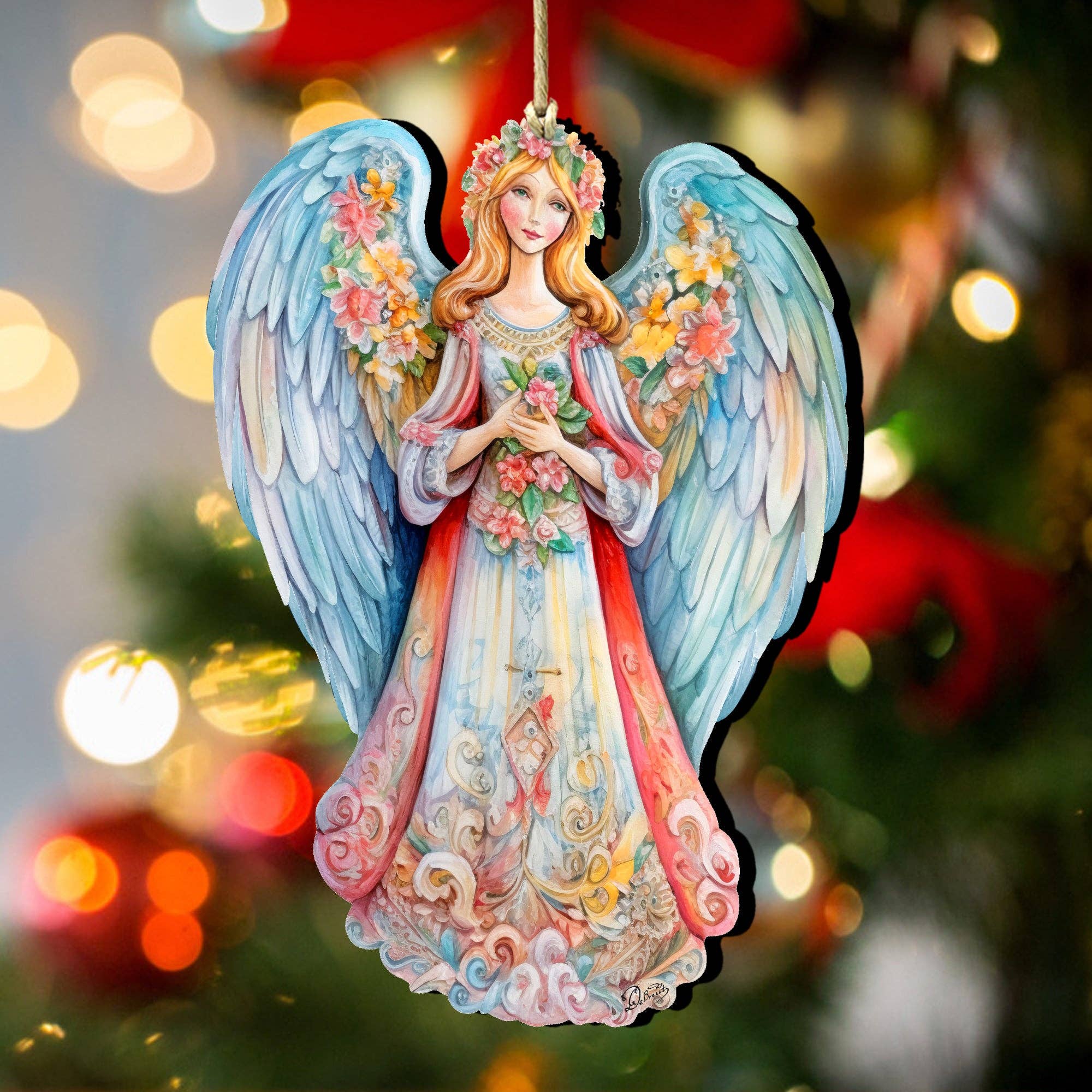 The "Ornament: Angel Blue Floral Wooden Ornaments Debrekht Nativity Holiday," a handcrafted keepsake with intricate wings and floral details, is displayed in front of a blurred, lit Christmas tree.