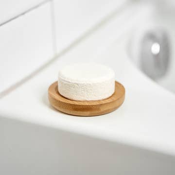 White round soap bar on a bamboo soap lift at the edge of a white sink, with tiled walls and a silver faucet in the background.