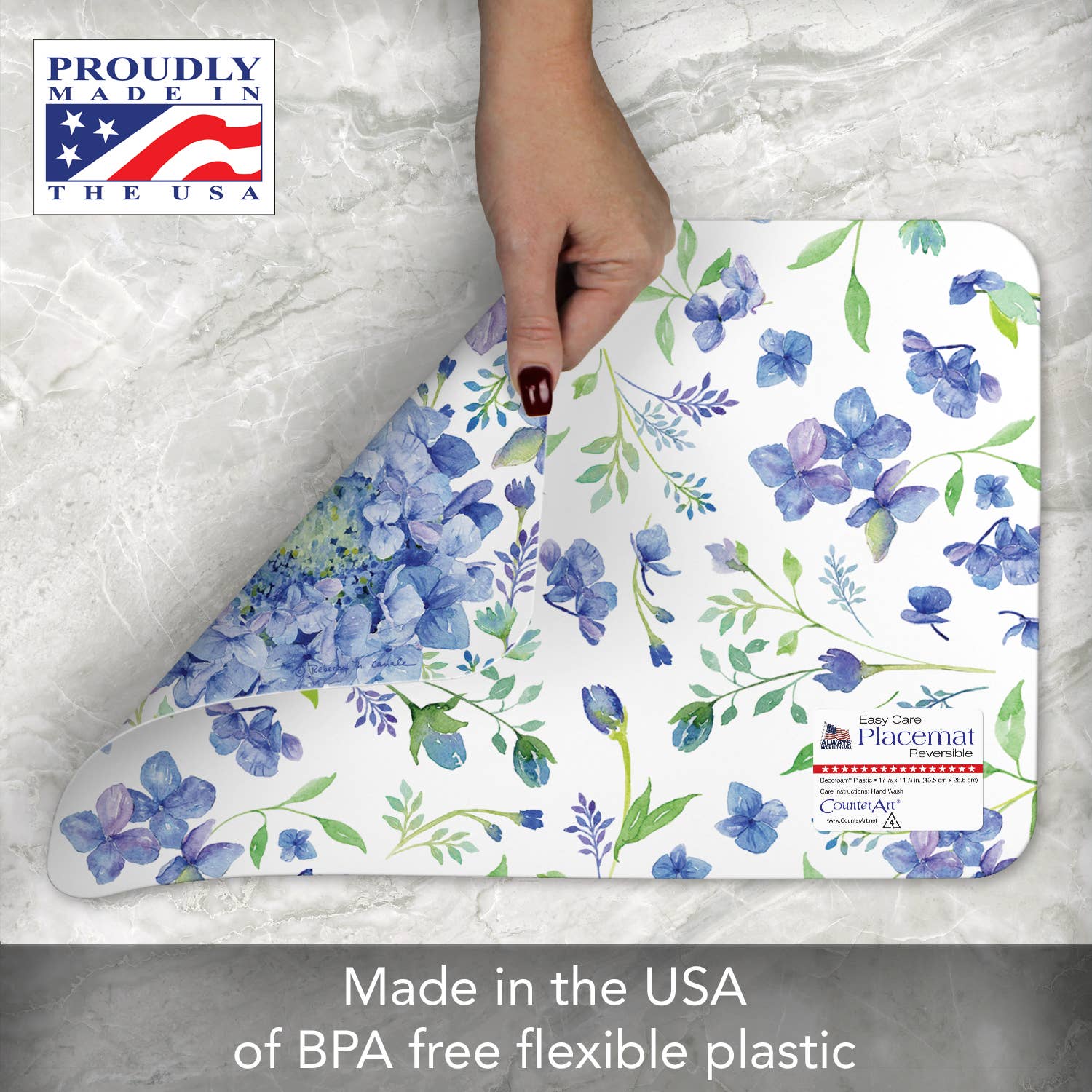 A hand lifts the Beautiful Hydrangea Reversible Rectangular Placemat on a marble surface. Text reads: Proudly Made in the USA and crafted from easy-care, BPA-free flexible plastic.