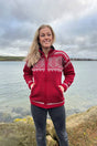 A woman standing by water wears a red and white zip-up wool sweater with a high collar and a Nordic pattern.