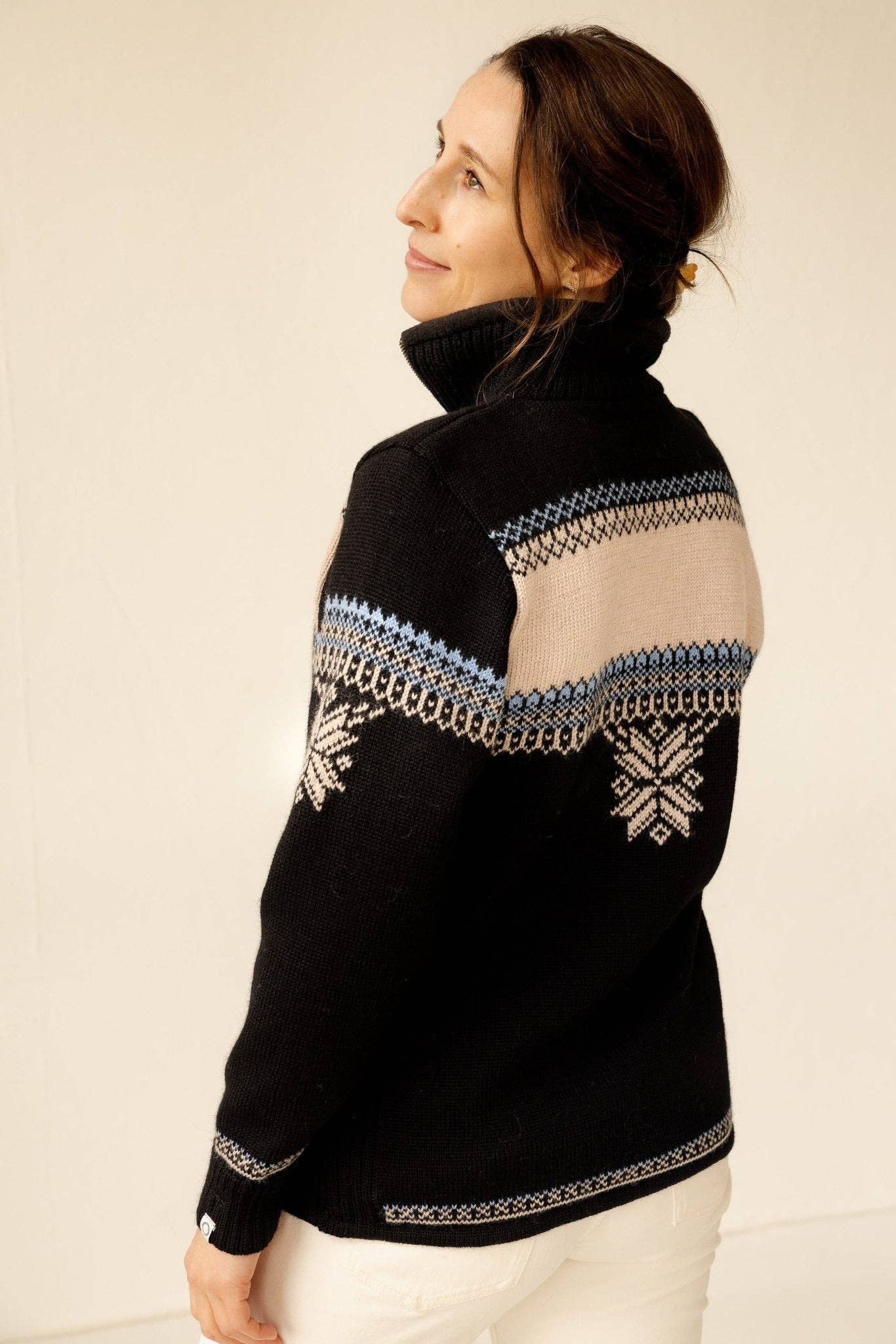 A woman in a black Voss Women's Sweater with beige and blue Nordic patterns stands with her back turned, looking over her shoulder.