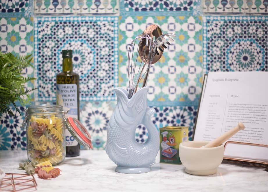 A charming kitchen counter features a fish-shaped utensil holder and the Gluggle Jug in white, set against a colorful tiled backsplash. A bottle of olive oil, jar of pasta, open cookbook, and mortar and pestle complete this whimsical culinary space.