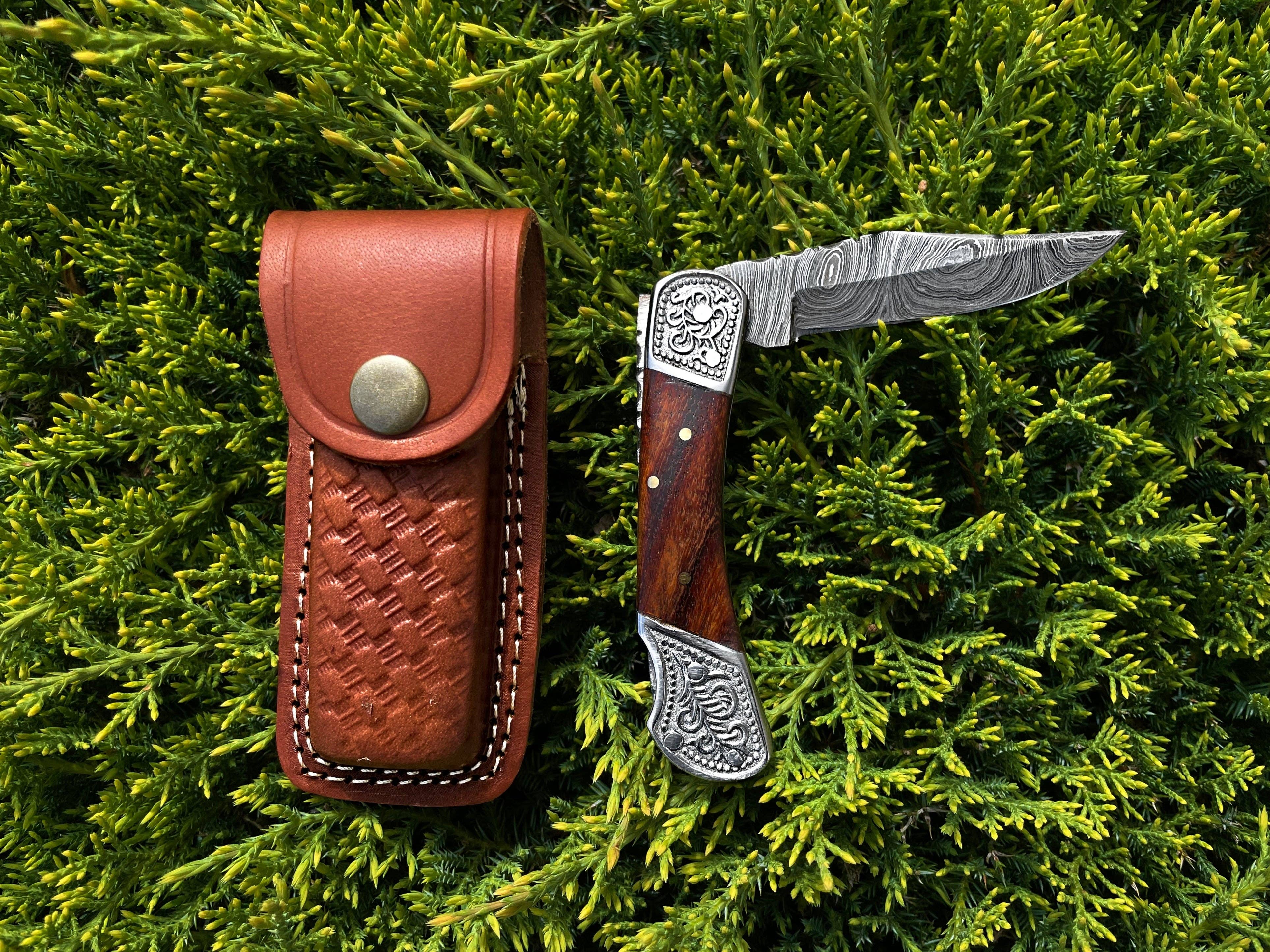 The Pocket Knife Walnut Wood 6.5'' Damascus Steel, featuring engraved metal accents and a walnut handle, is displayed near its leather sheath on green foliage.