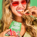 Woman with long blonde hair wearing pink sunglasses and a light green top, holding a Swedish gummy candy bag labeled “Bites by Sweden Sour Mix” and eating a gummy.