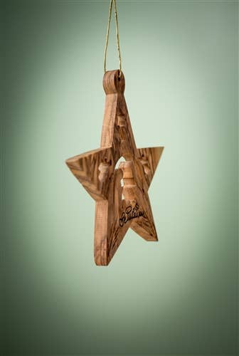 The Ornament: Star with Nativity and Angels is a 3" Christmas tree ornament from Bethlehem, featuring a carved scene, hanging from a gold string against a green background.