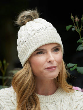 A woman outdoors with greenery behind her is wearing the Chunky Cable Hat - Natural (One Size), a cream-colored cable knit hat featuring a faux fur pom-pom, along with a matching sweater.
