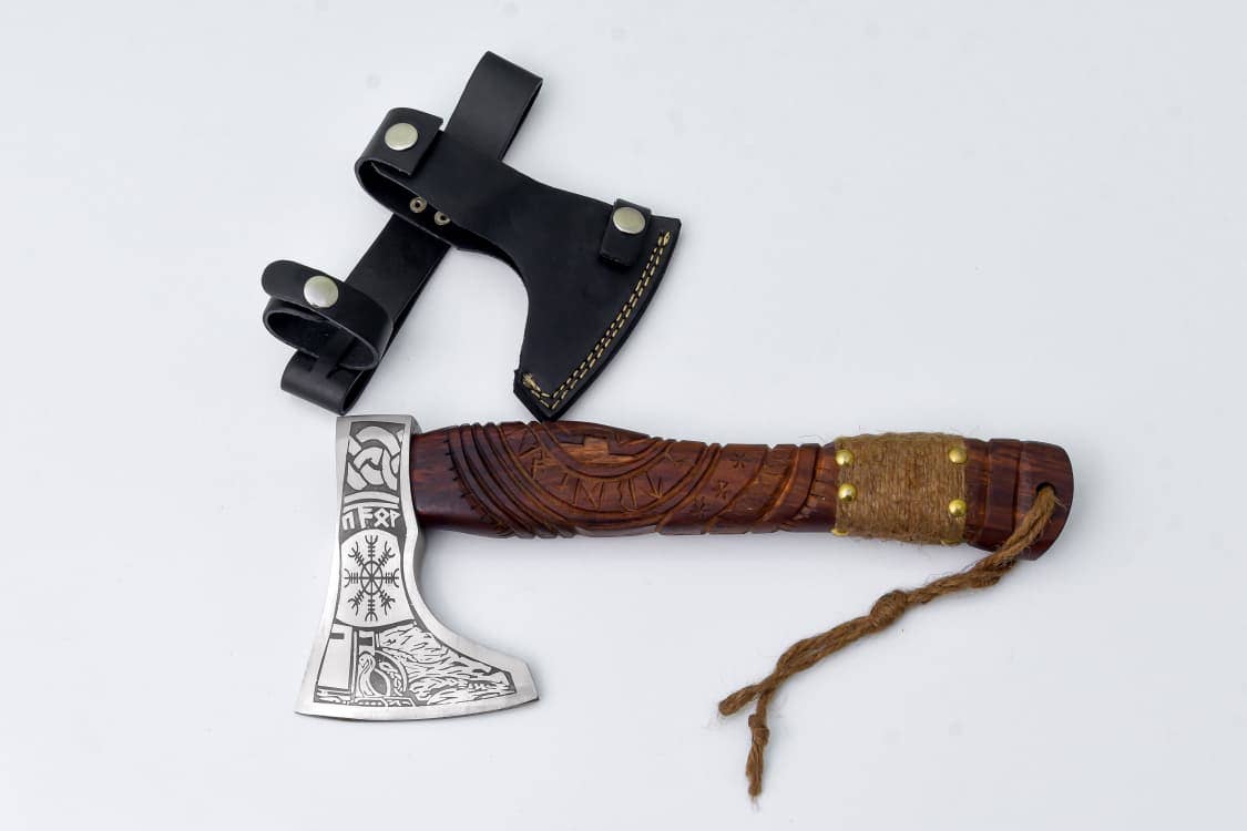 The Viking Axe "Fenrir's Mark" is a handcrafted, engraved carbon steel hatchet with a runic carved handle, pictured beside its black leather sheath with snap closures on a white background.