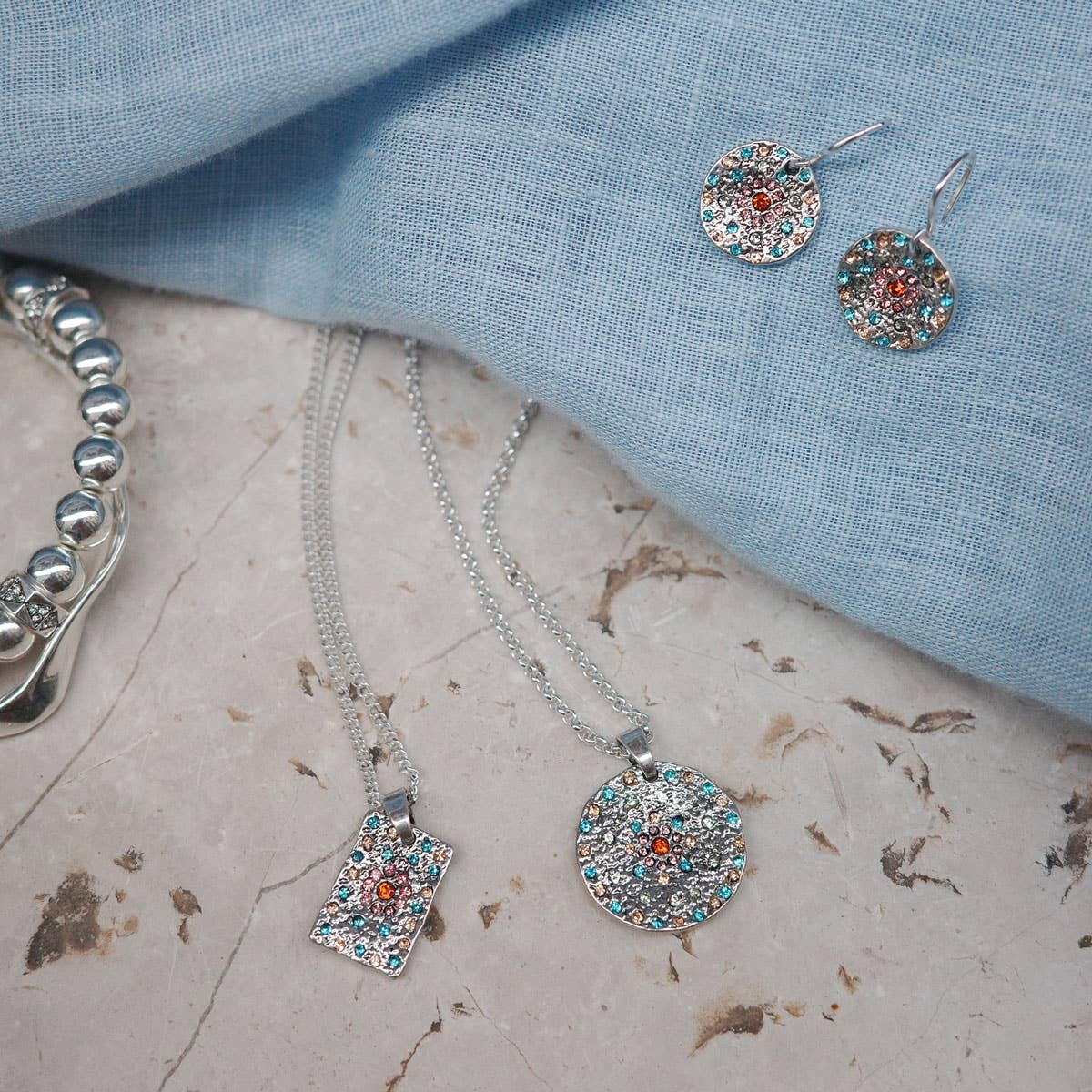 The jewelry set includes the A&C Oslo Vibrance Silver Coins Earrings, a necklace, and a bracelet adorned with round and rectangular pendants, all elegantly displayed on gray stone and light blue fabric.