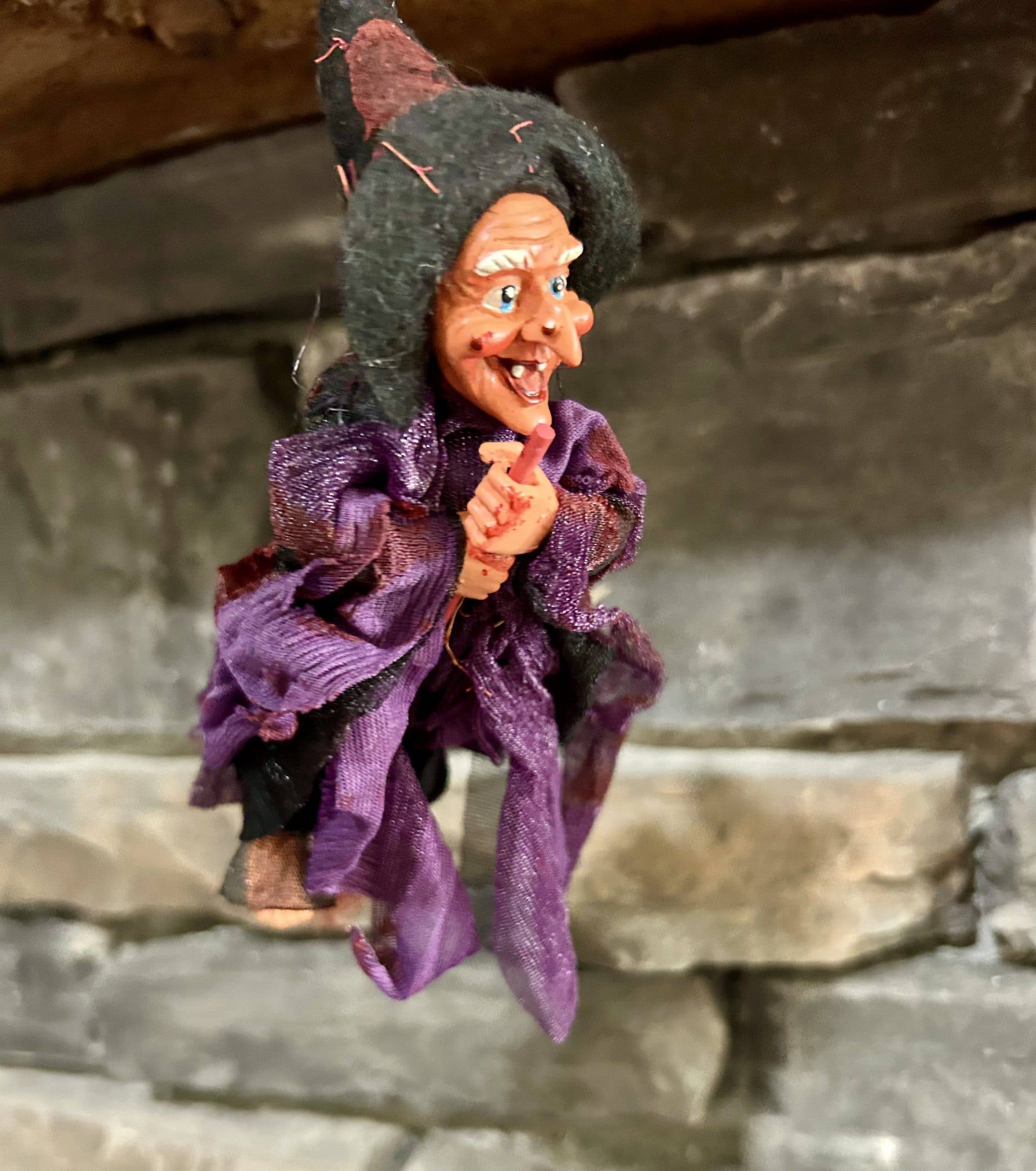 The Walburga Purple & Black Kitchen Witch Doll figurine, 8" tall, hangs in front of a stone wall—an enchanting good luck charm for any home.