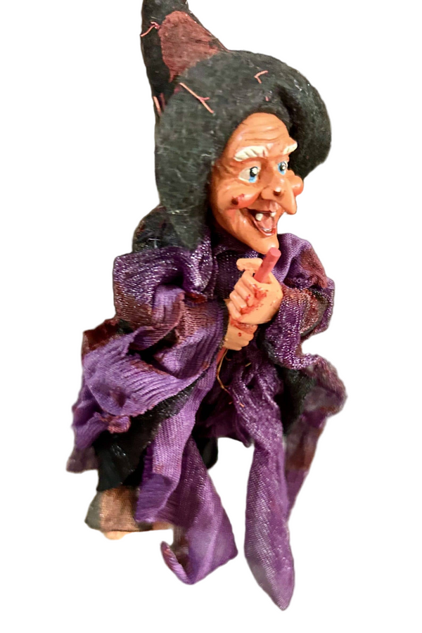 The Walburga Purple Kitchen Witch Doll Figurine is an 8" tall hanging broom rider, dressed in a purple dress and black hat—complete with exaggerated features and a mischievous grin for kitchen protection or good luck.