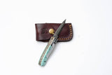 The Titan International Turquoise Damascus Folding Knife – Hand Engraved Bolster Edition features a turquoise inlay and silver handle, resting on its brown leather sheath with tan stitching and brass button, set against a white background.