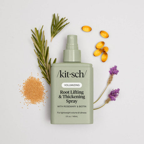 A bottle of Hair: Root Lifting & Thickening Spray 5 fl oz by Kitsch is displayed with rosemary, lavender, amber capsules, and granules on a light background, emphasizing this volumizing spray's botanical ingredients.