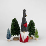 A wool Tomte gnome with a tall gray hat and red body stands upright among four small artificial trees on a plain white background.