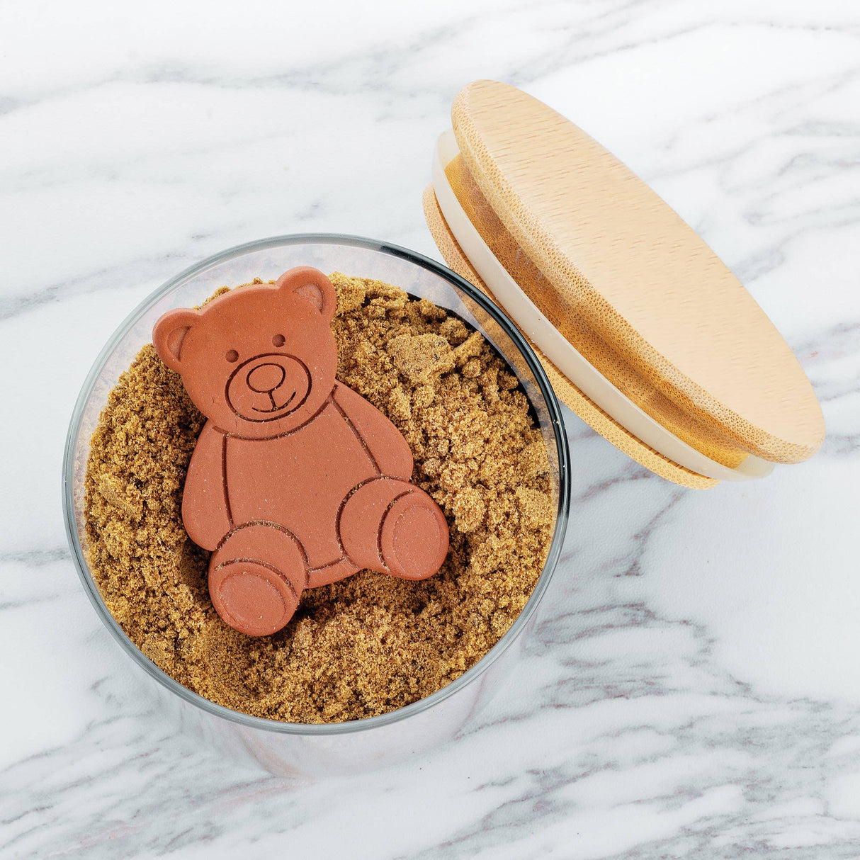The Sugar Saver: Terracotta Brown Sugar Bear Keeper & Saver, 2.5", sits on brown sugar in a glass jar with a wooden lid nearby on a marble surface.