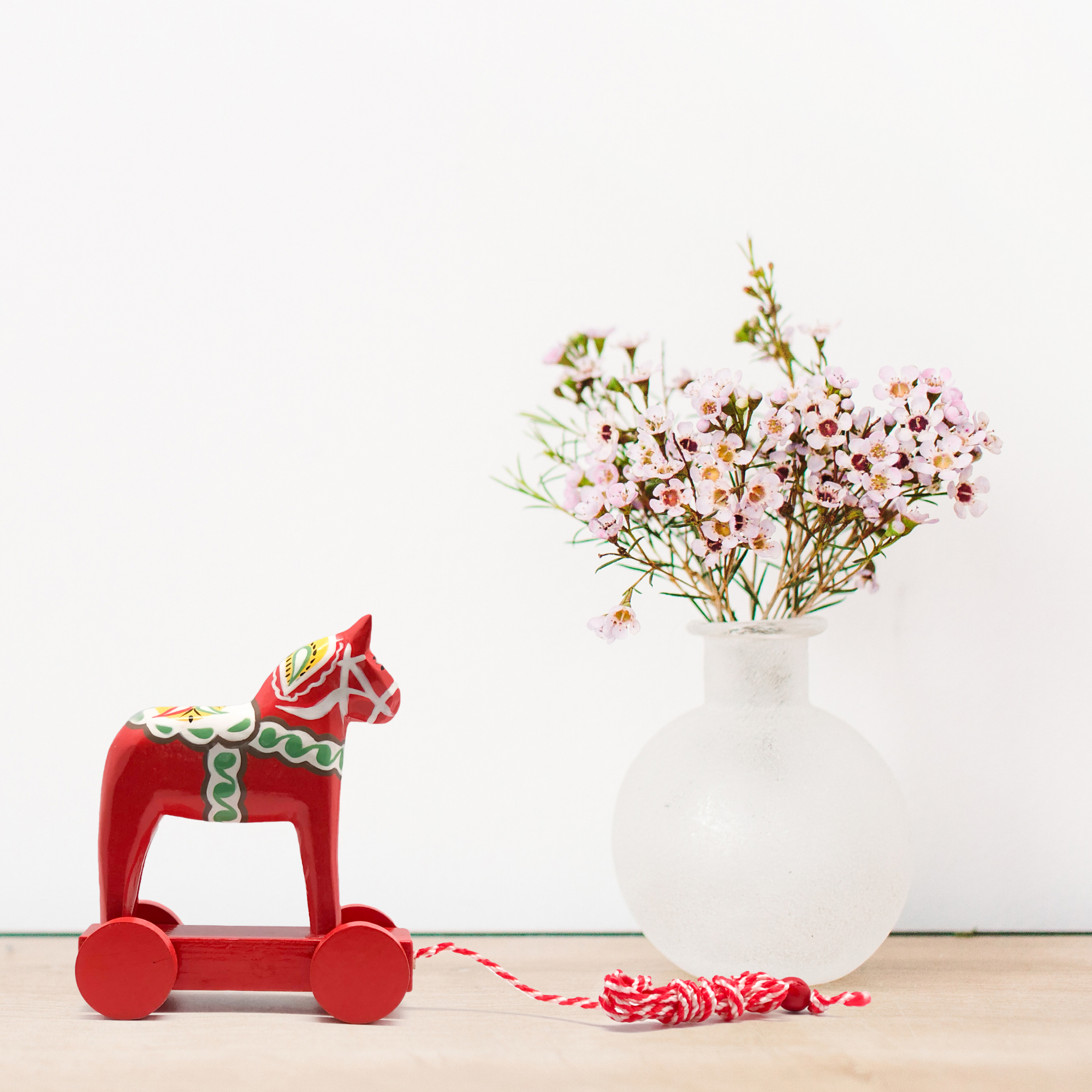 A Swedish Horse Themed Pull Toy Dala Horse adds a touch of Scandinavian charm as it rests beside a frosted vase filled with small pink flowers on a wooden surface.