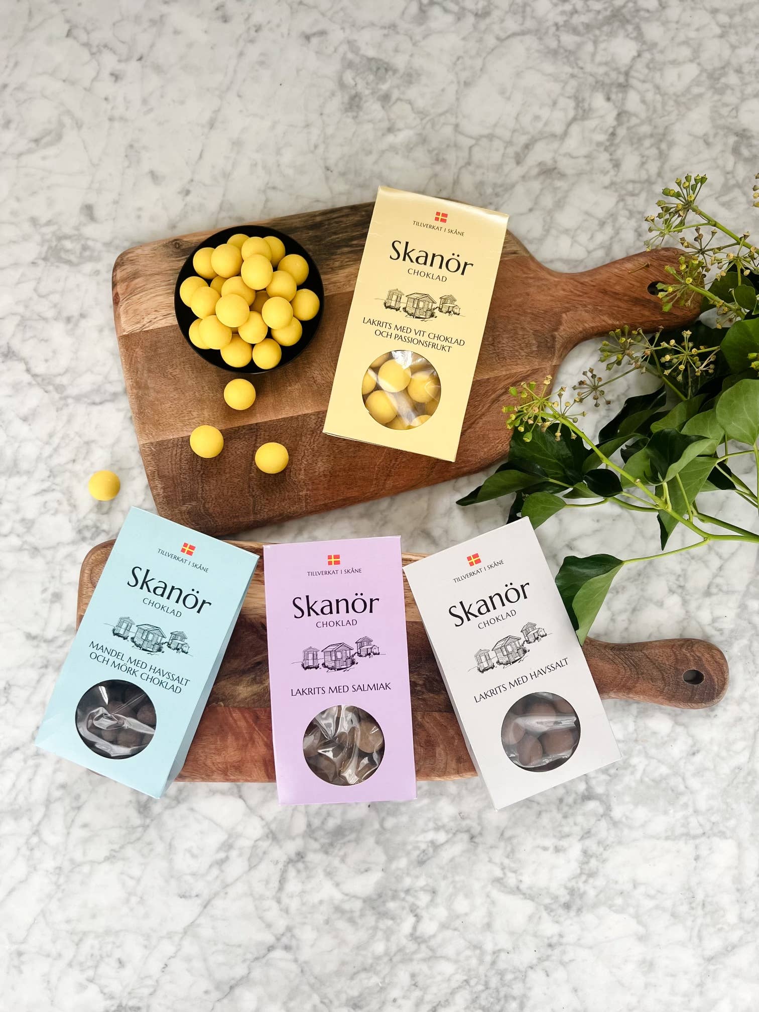 Boxes of Skanör Chocolate Coated Licorice with Sea Salt from Skåne, Sweden, grace a wooden board with scattered yellow candies, surrounded by green leaves.