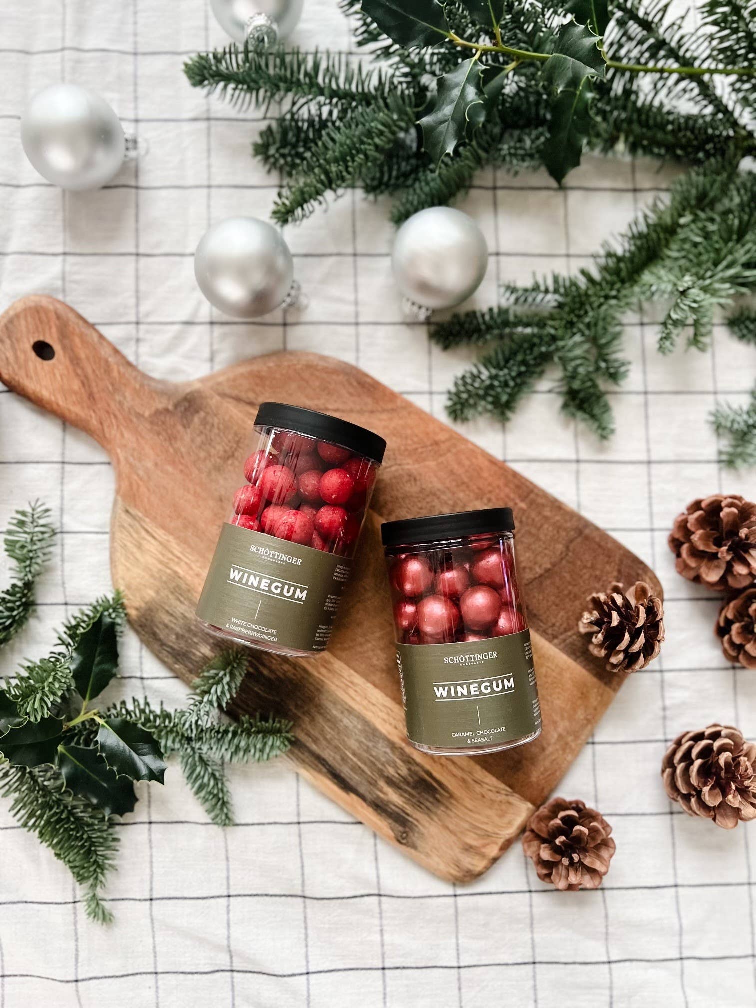 Two jars of Candy: Classic Winegum Seasalt & Caramel Chocolate 250 g rest on a wooden cutting board, surrounded by pinecones, greenery, and silver ornaments on a checkered tablecloth.