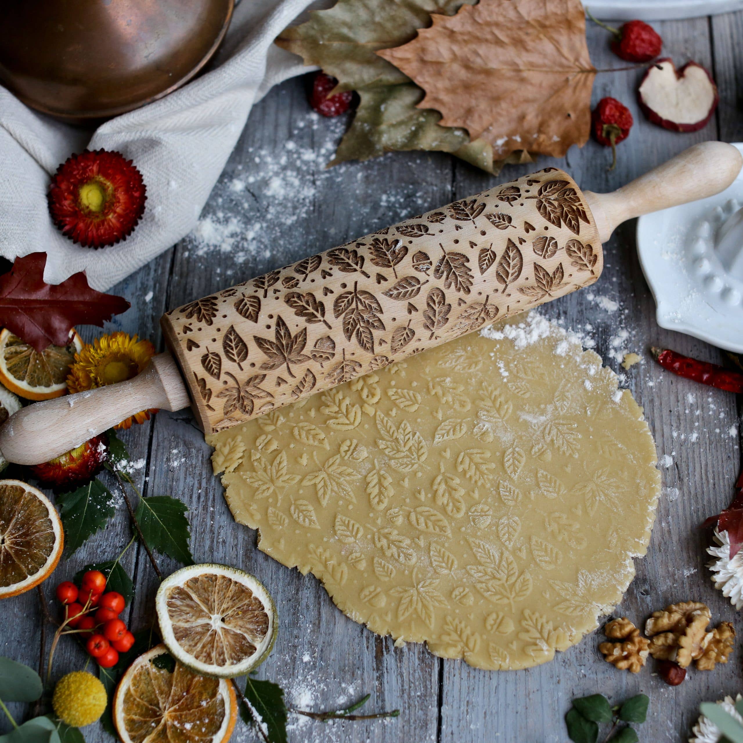 The Autumn Leaves Embossing Rolling Pin, surrounded by dried fruits, nuts, and autumn leaves on a rustic wooden surface, rolls across the dough with intricate leaf patterns.