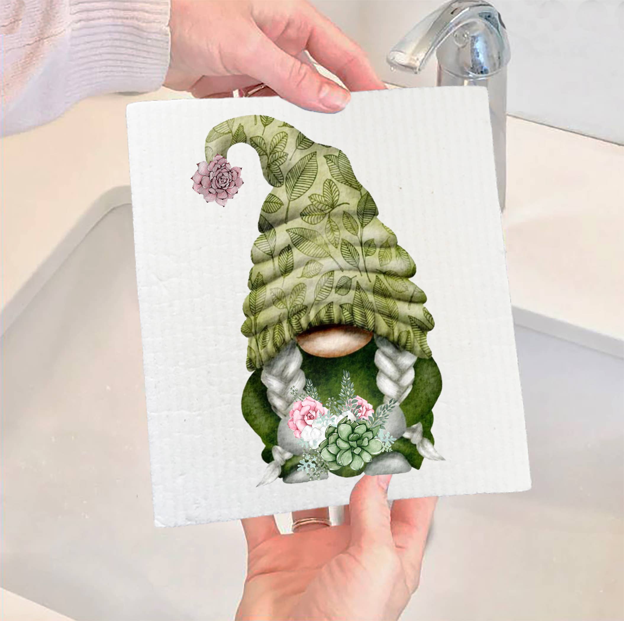 Hands hold the "Succulent Desert Green Gnome Swedish Dish Cloth," showcasing a whimsical design of a gnome with braids and a tall green hat embellished with leaves and flowers. This eco-friendly dishcloth is ideal for kitchens, as evidenced by the sink and faucet in the background.