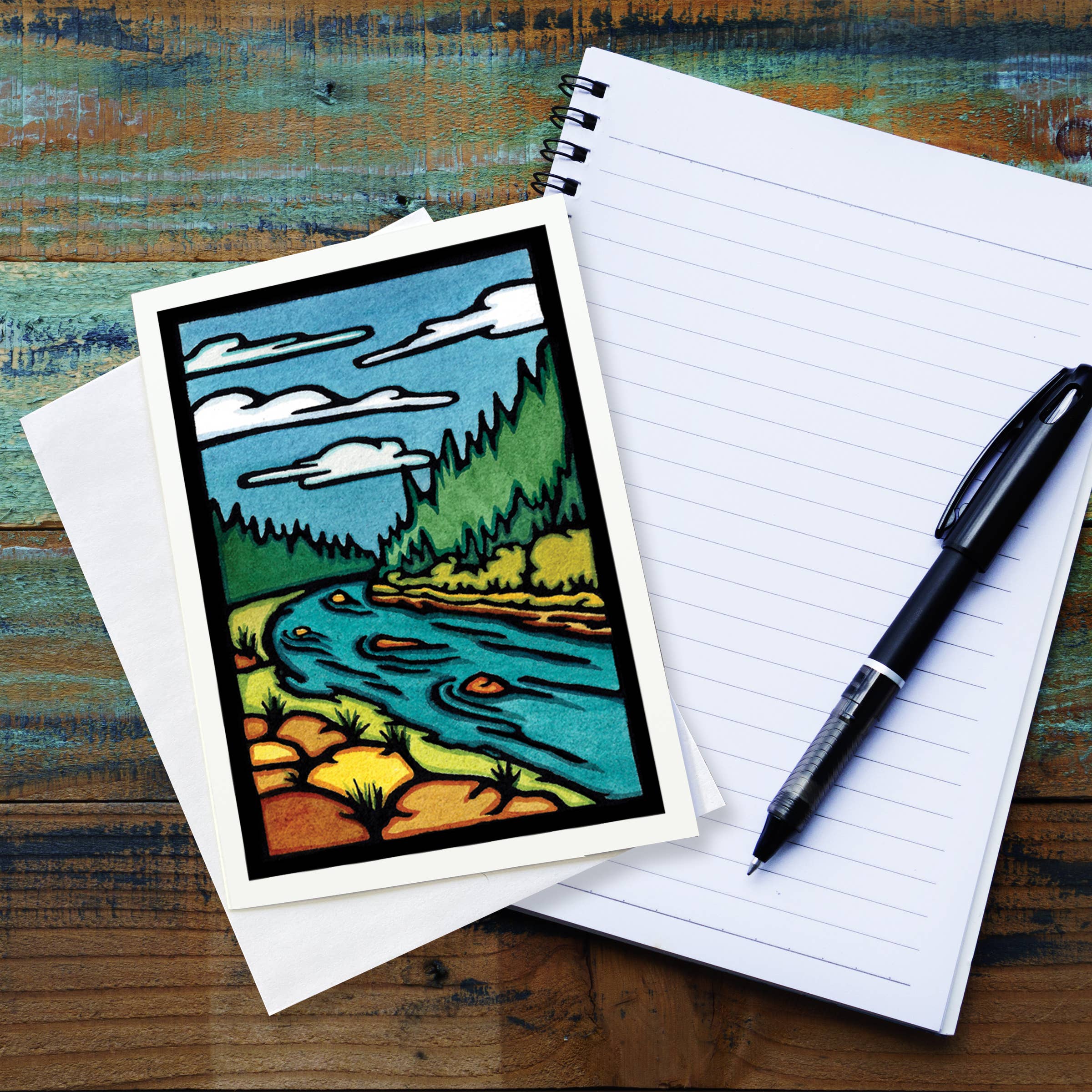 The vibrant "Greeting Card: The River" by Sarah Angst, showcasing a colorful landscape with a river and trees, rests on a lined notebook, accompanied by a black pen.