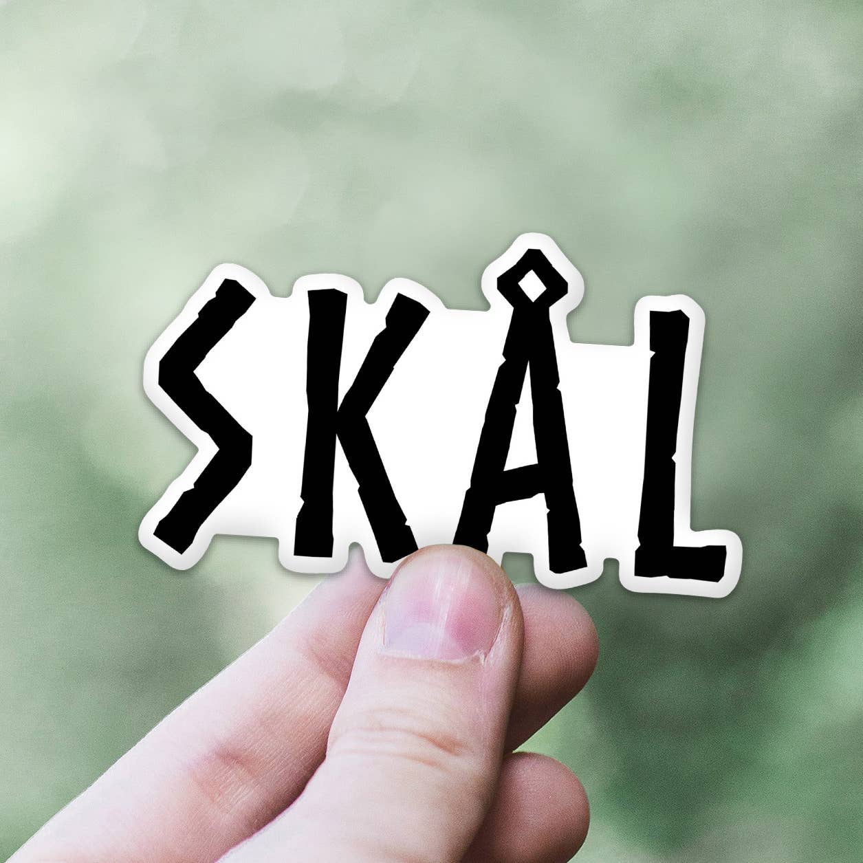 A hand holding a Sticker: Skål Viking Sticker that reads SKÅL in bold black letters with a blurred green background, celebrating its fade-resistant and weather-proof design.