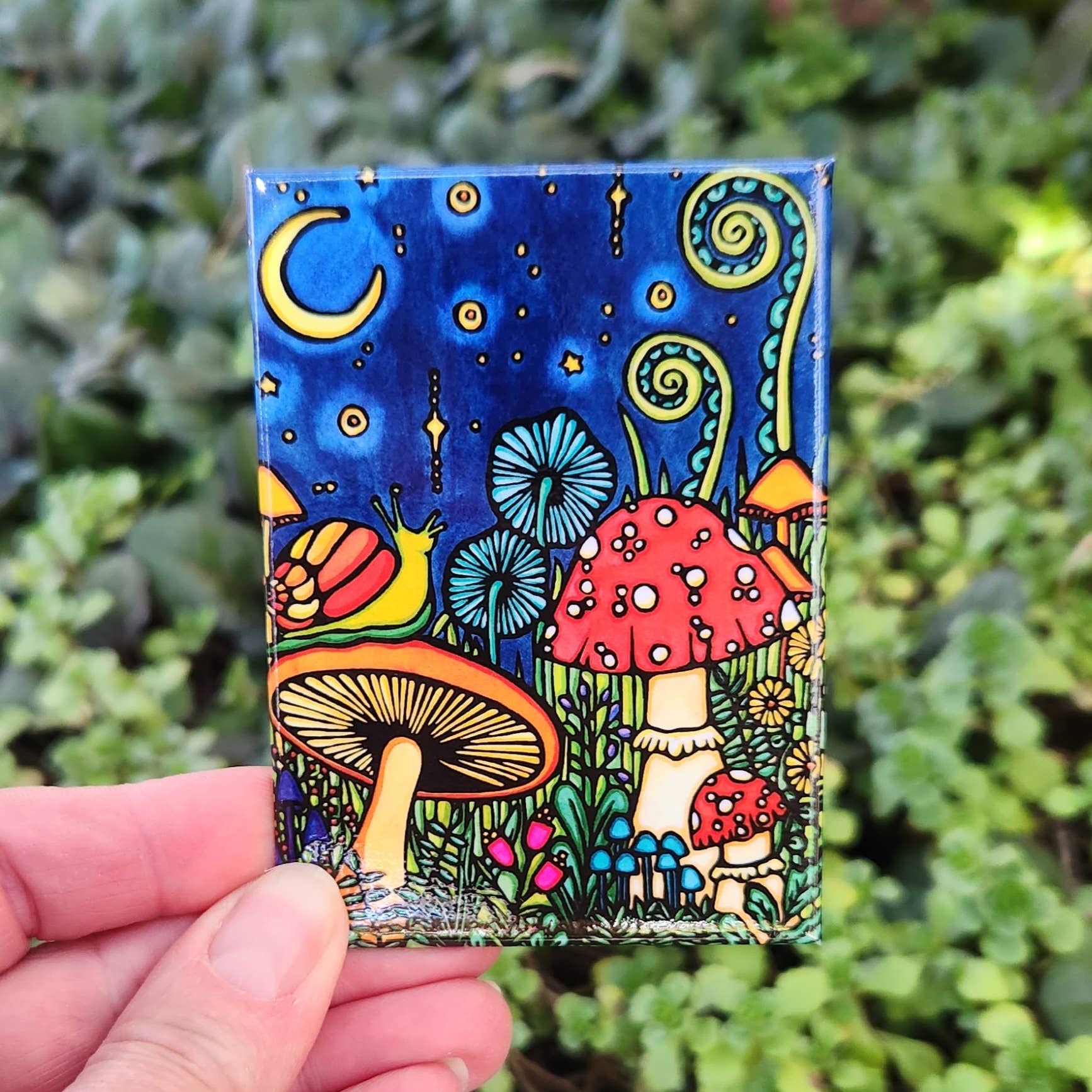 A hand holds the vibrant Mushroom Magnet, featuring mushrooms, plants, a snail under a crescent moon and starry night sky.