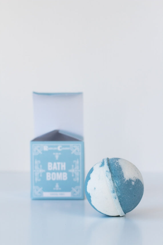 A blue and white Coastal Vibes Bath Bomb sits before a light blue box labeled BATH BOMB on a white background. Each handmade bath bomb is crafted in Virginia Beach for a luxurious, skin-nourishing bath experience.